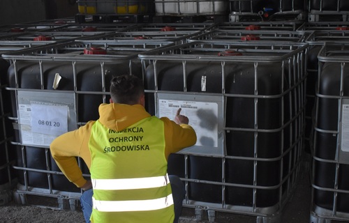 Inspektor Wojewódzkiego Inspektoratu Ochrony Środowiska w Warszawie przeprowadza oględziny miejsca porzucenia odpadów. W tle pojemniki o pojemności 1000 l w hali magazynowej.