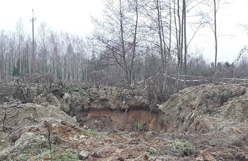 Wykop w ziemi o powierzchni kilku metrów kwadratowych na otwartym terenie, wcześniej przygotowany do zakopania odpadów, które zostały usunięte. W tle las.